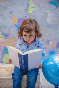 girl reading book while sitting beside desk globe